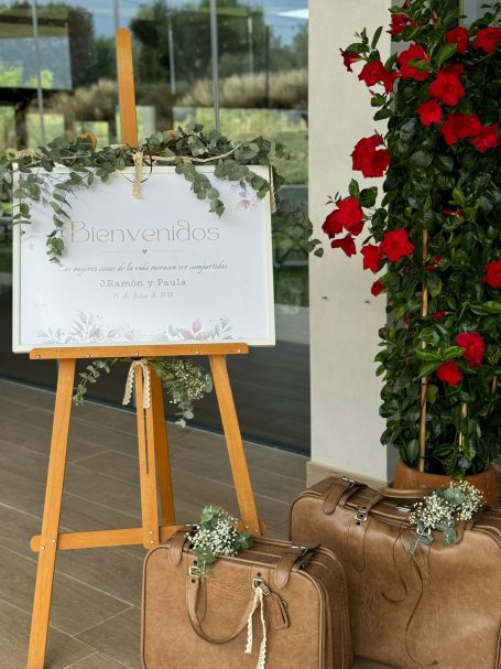 Cartel de bienvenida decorado con flores junto a maletas en un entorno elegante.